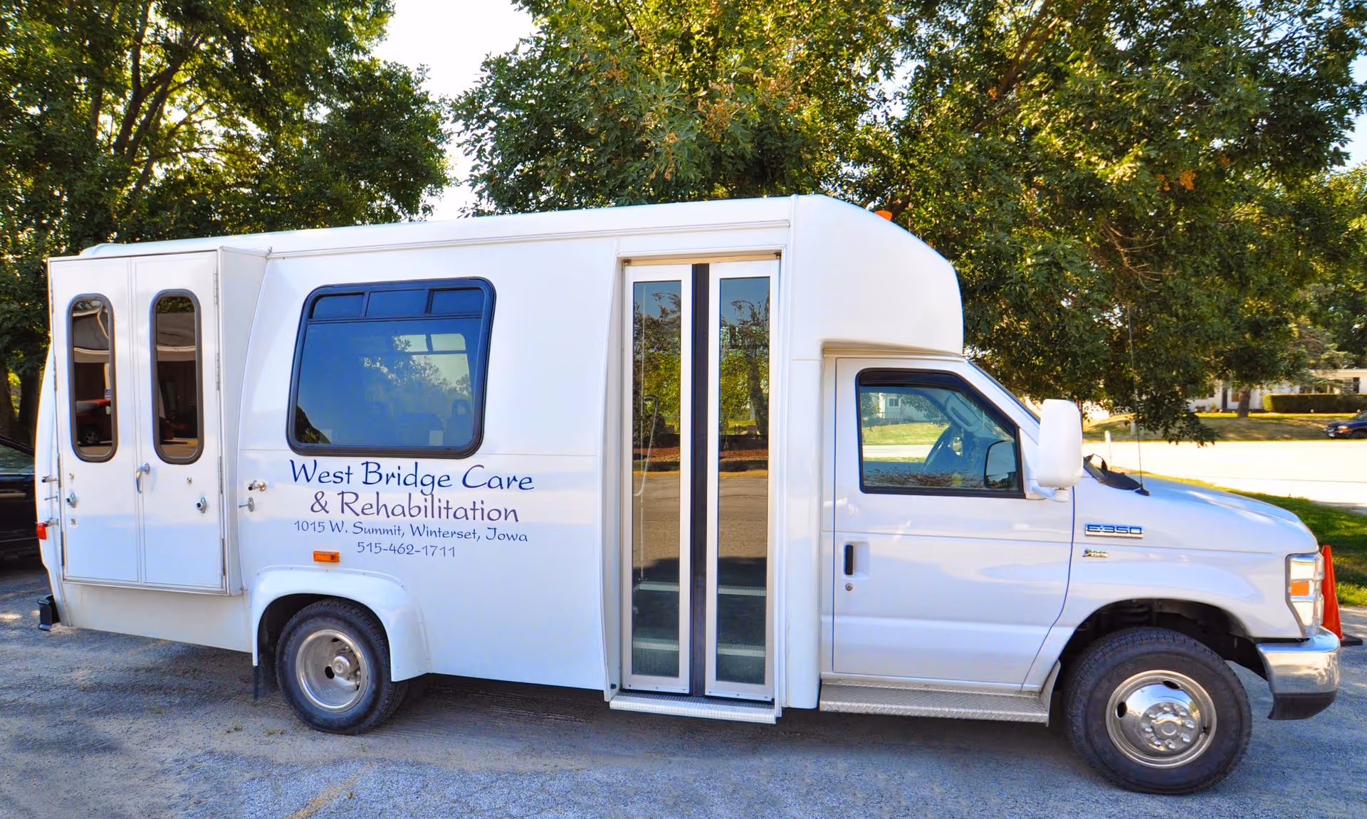 A white shuttle van parked outdoors with the text 'West Bridge Care & Rehabilitation, 1015 W. Summit, Winterset, Iowa, 515-462-1711' on the side. The van has large windows and a sliding door, with trees and a street visible in the background.