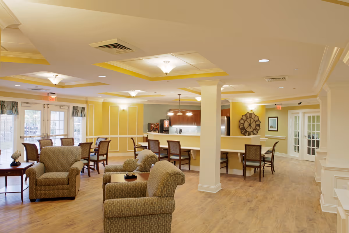 A spacious and well-lit common area in a senior living facility featuring comfortable armchairs, several dining tables with chairs, a kitchen counter with bar stools, and decorative wall elements including a large clock. The room has light yellow walls, wooden flooring, and ceiling lights with recessed panels. French doors and large windows allow natural light to enter the space.