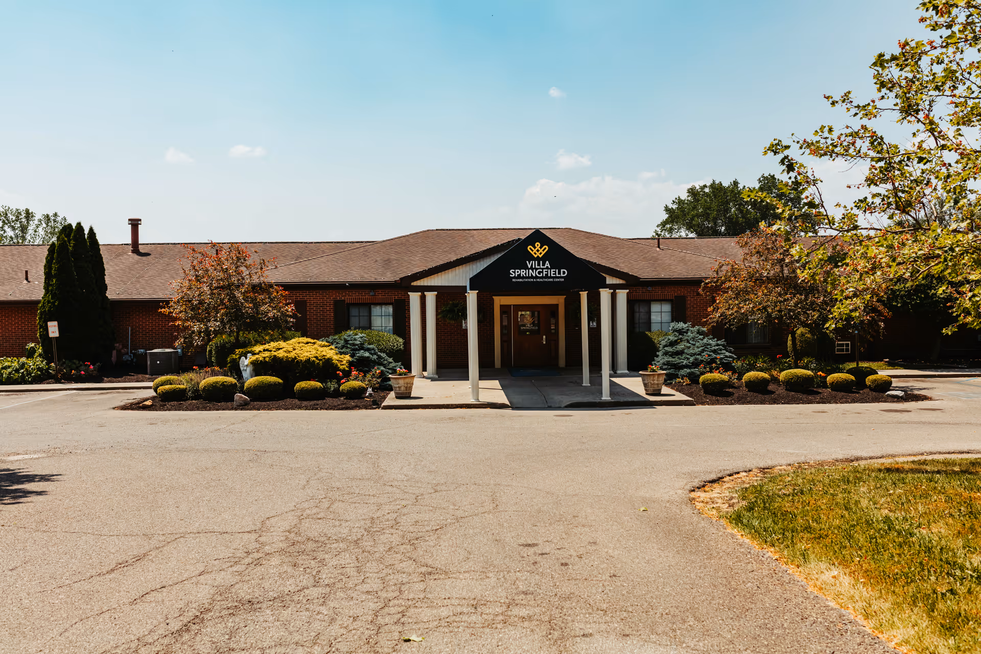Front exterior view of Villa Springfield Rehabilitation and Healthcare Center, a single-story brick building with a covered entrance supported by white columns, surrounded by landscaped bushes and trees under a clear blue sky.