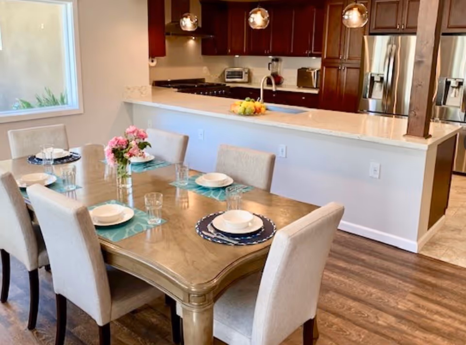 A dining area with a wooden table set for six people with plates, bowls, glasses, and placemats. A vase with pink flowers is in the center of the table. Behind the dining area is a kitchen with dark wood cabinets, a white countertop, stainless steel refrigerator, and hanging pendant lights.