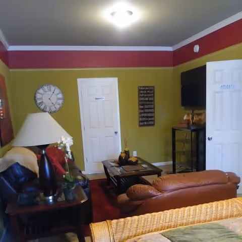 A cozy living room with green and red walls, featuring a black leather couch with a beige throw, a brown leather recliner, a wooden coffee table with decorative items, a side table with a lamp and a plant, a wall clock, a wall-mounted TV, and two white doors.