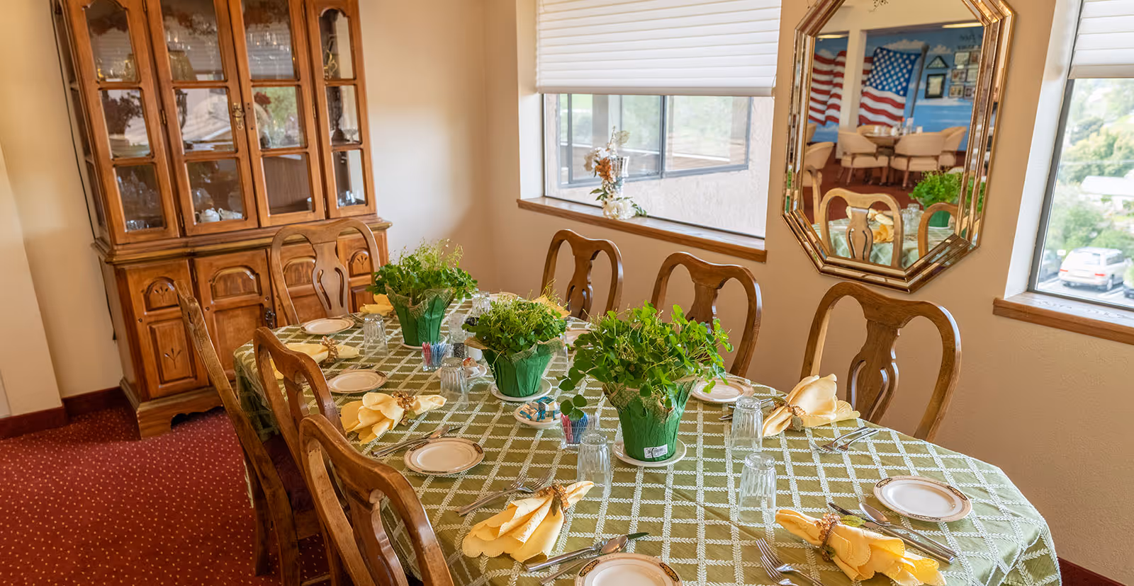 A dining room with a long wooden table covered with a green checkered tablecloth. The table is set with plates, glasses, silverware, and yellow napkins. There are three green potted plants as centerpieces. Wooden chairs surround the table. A wooden china cabinet with glass doors is against the wall. A large mirror on the wall reflects another dining area with American flag decorations. Two windows provide natural light and a view of the outside parking area.