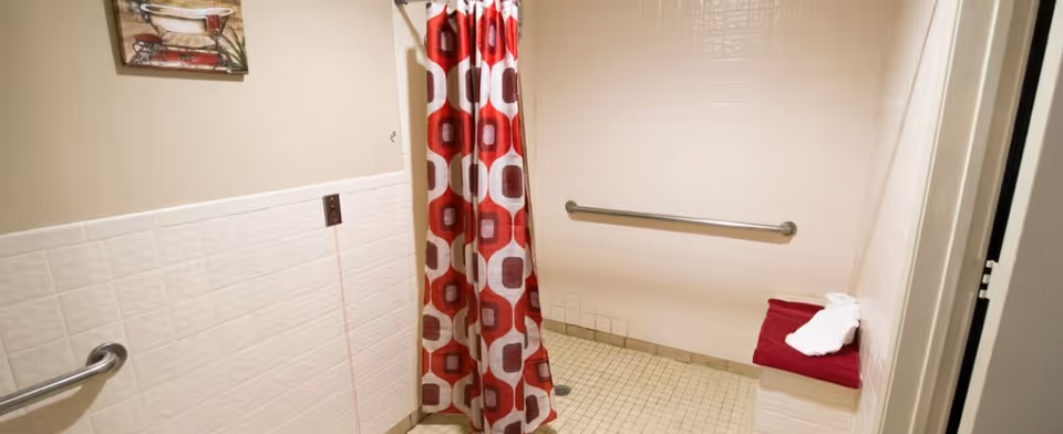 A tiled shower area with a red and white patterned shower curtain, grab bars on the walls, and a small bench with a folded white towel on a red cloth. The walls are partially tiled in white, and there is a small painting of a bathtub hanging on the wall.