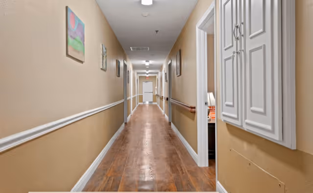 A long, well-lit interior hallway with wood flooring, beige walls, handrails, artwork, and doorways to resident rooms.