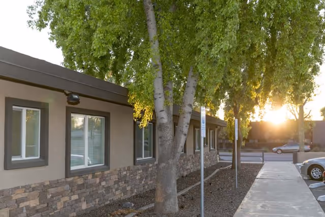 Exterior view of Desert Terrace Healthcare Center showing a building with stone and stucco facade, several windows, a sidewalk, trees with green leaves, and a parking area with cars at sunset.