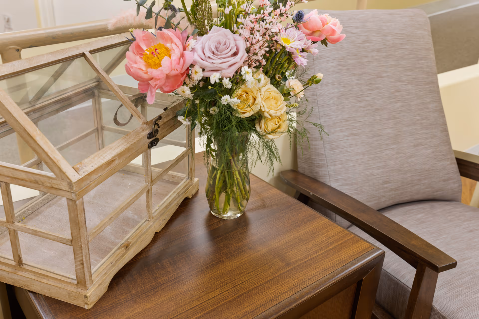 A vase of mixed flowers and a small wooden-and-glass terrarium sit on a wooden side table beside a beige upholstered armchair.