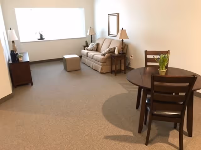 A cozy living room area with a beige sofa, two side tables with lamps, a small ottoman, a wooden dining table with three chairs, and a large window letting in natural light. The walls are plain white and the floor is carpeted.