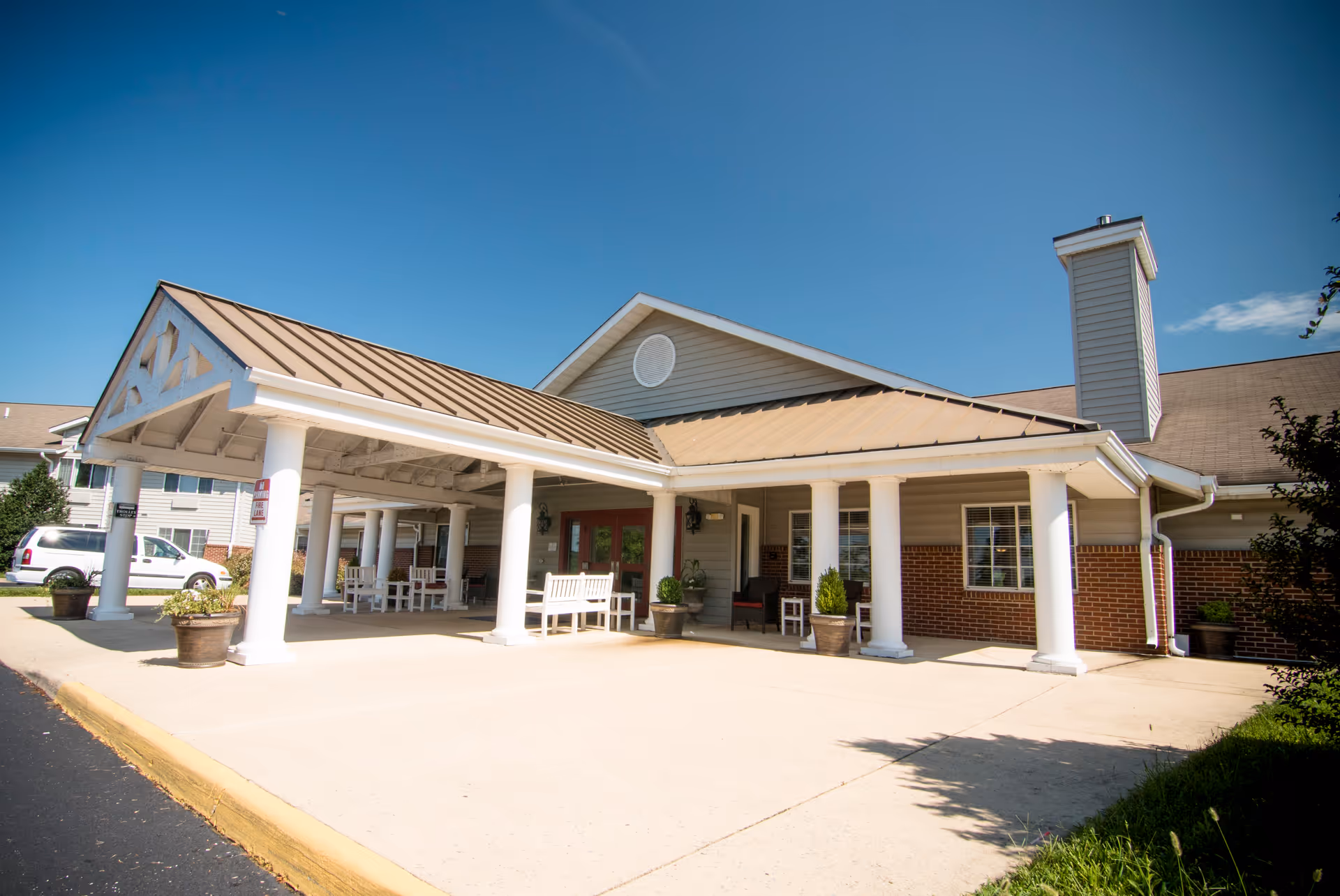 Exterior view of Brookdale Staunton facility entrance with a covered drop-off area supported by white columns, benches, potted plants, and a clear blue sky.