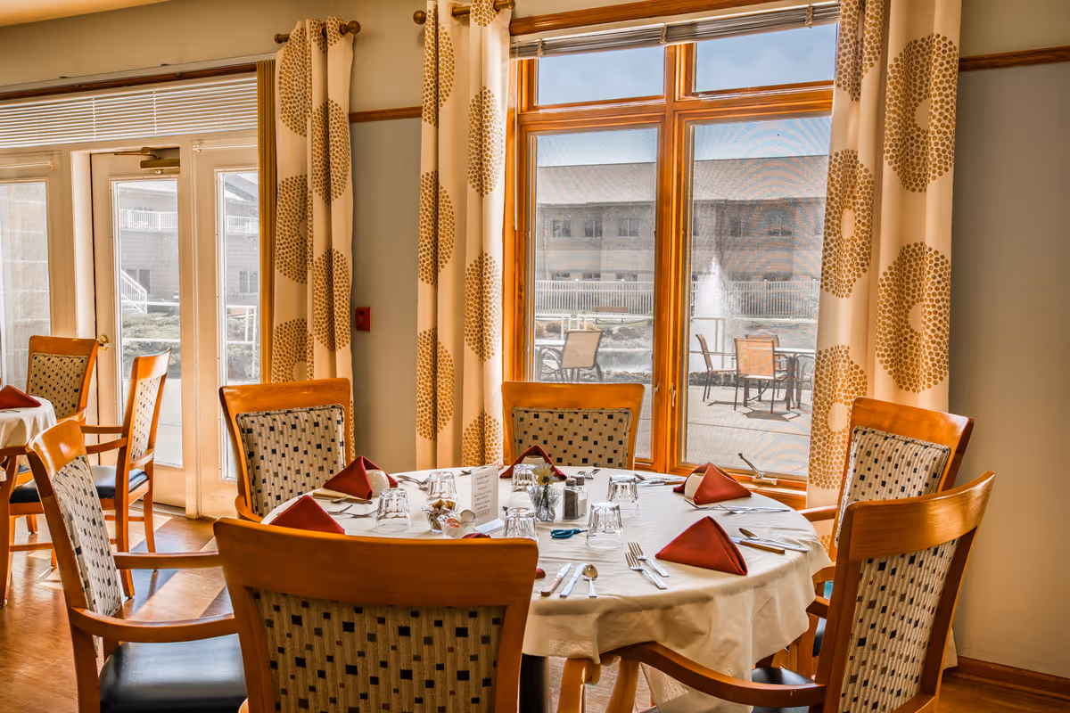 A dining room with a round table set for six people. The table is covered with a white tablecloth and has red folded napkins, glasses, and silverware arranged neatly. The room has large windows with patterned curtains, allowing natural light to brighten the space. Outside the windows, there is a patio area with chairs and a water fountain.
