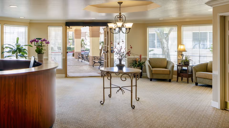 A bright and welcoming senior living facility lobby with a curved wooden reception desk on the left, a round table with a flower arrangement in the center, and two armchairs with a side table and lamp near large windows on the right. The entrance door is open, showing an outdoor patio area with benches and columns.