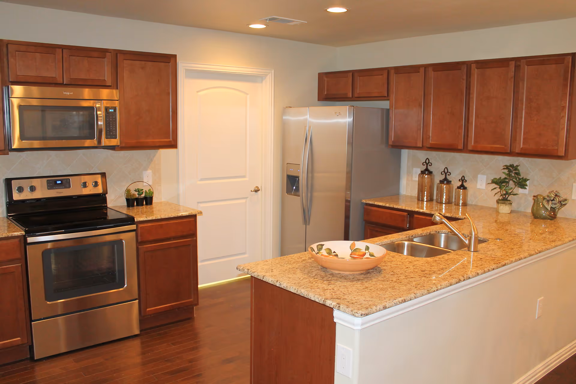 Modern kitchen with wooden cabinets, stainless steel appliances including a microwave, oven, and refrigerator. Granite countertops with a double sink and decorative items such as a bowl, plants, and jars are visible. A white door is in the background.