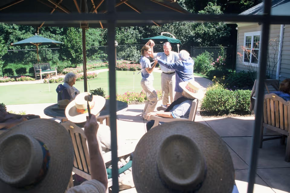 A group of elderly people sitting and socializing outdoors on a patio with umbrellas and garden furniture. In the background, a caregiver is dancing with two elderly individuals near a well-maintained garden and putting green area.