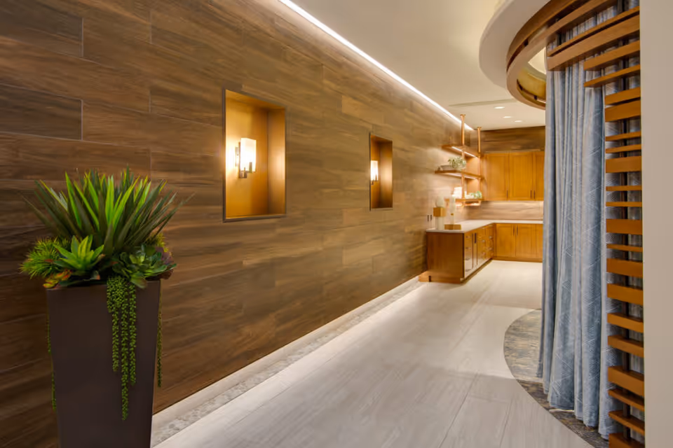 Well-lit interior corridor with a wood-paneled wall, recessed sconces, a tall planter, and a small kitchenette and curtained seating nook.