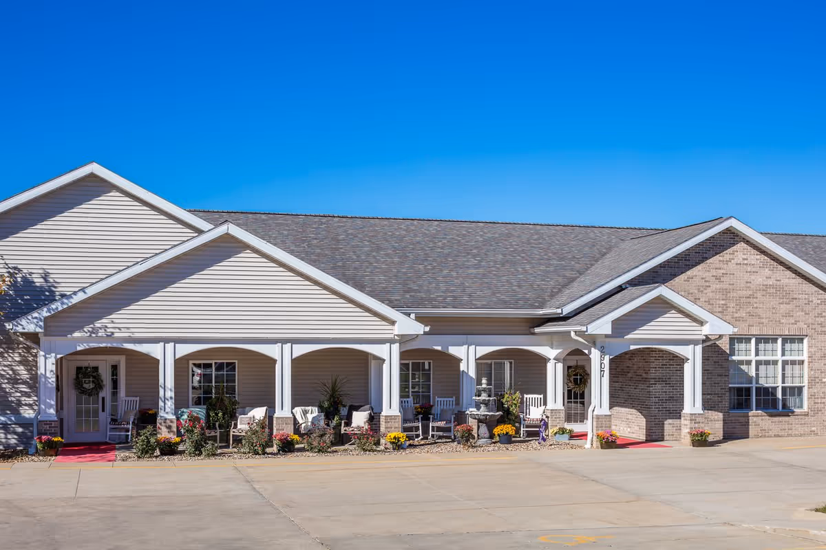 Exterior front view of Glenwood Place facility showing a single-story building with beige siding and brick accents. The entrance features a covered porch with white columns, rocking chairs, potted plants, flowers, and a decorative fountain. The sky is clear and blue.