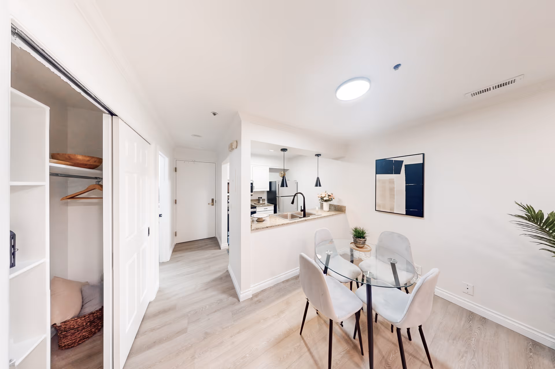 Bright apartment dining area with a round glass table and four chairs next to a pass-through kitchen and an open closet.