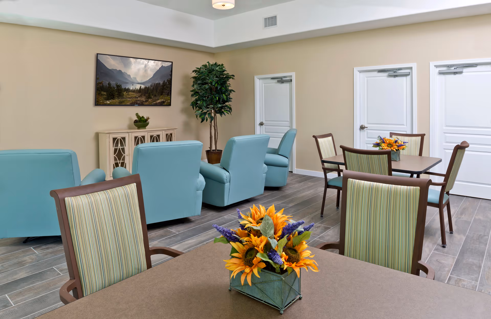 A common area in an assisted living facility featuring a table with a floral centerpiece in the foreground, several chairs with striped upholstery, and a seating area with light blue armchairs facing a wall-mounted landscape painting. There is a potted plant near two white doors on a beige wall and wood-look tile flooring.