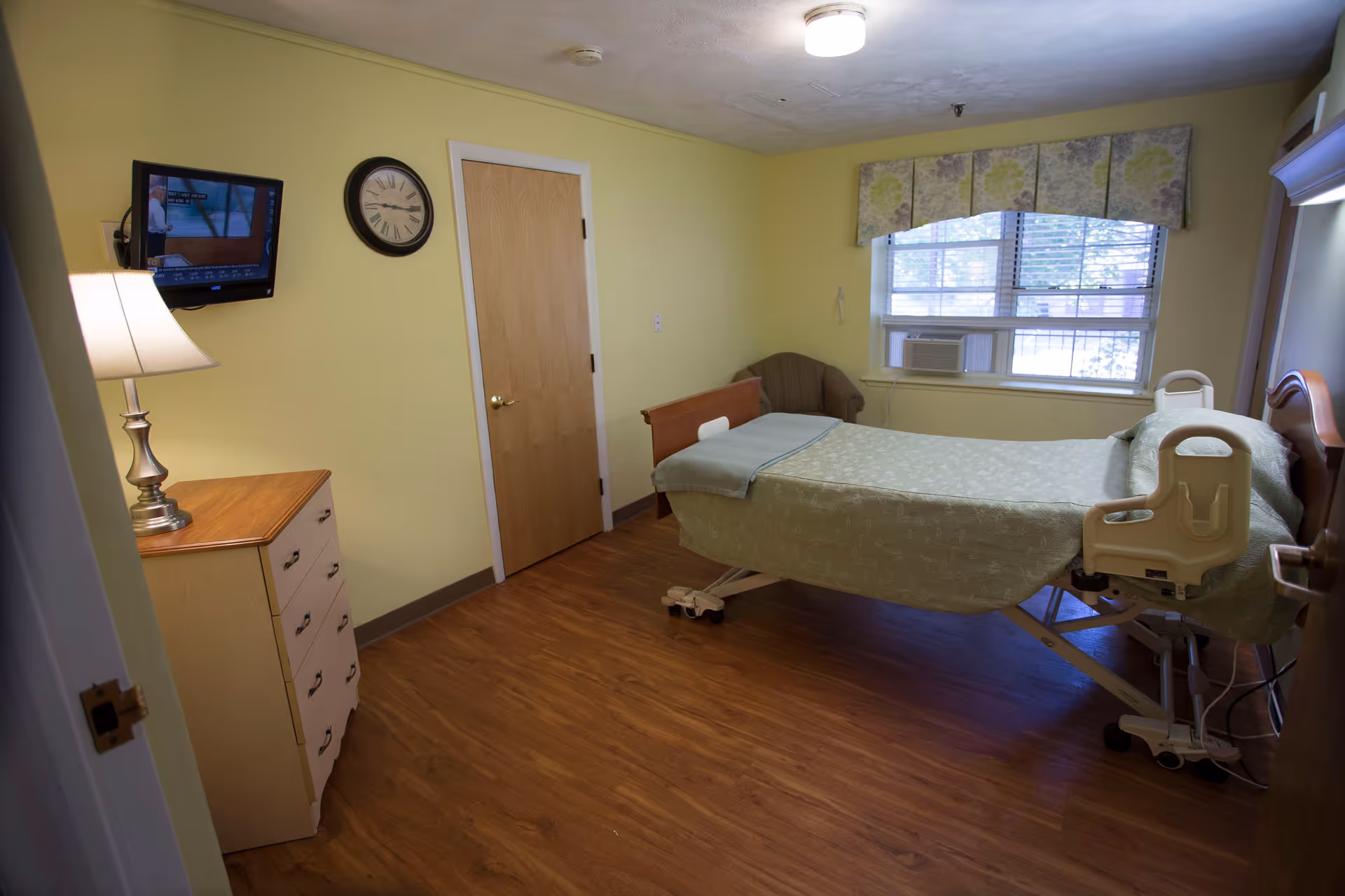 A small senior facility private bedroom with an adjustable bed, dresser, wall clock and TV under a window with blinds.