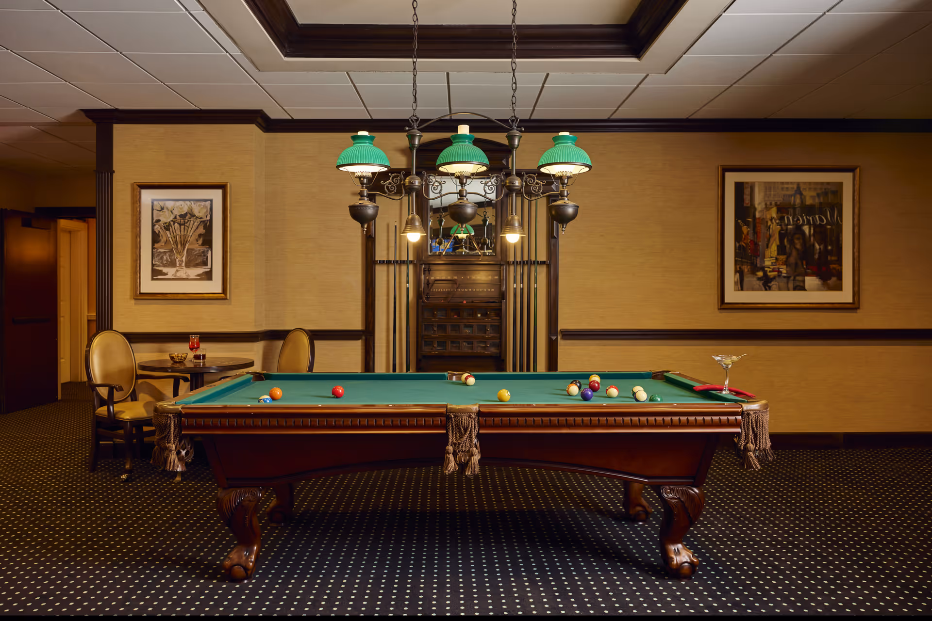 A classic billiards room with a green felt pool table in the center, surrounded by a dark wood frame and ornate legs. Above the table hangs a vintage-style light fixture with green glass shades. Behind the table is a rack holding pool cues and a mirror. To the left, there is a small round table with two upholstered chairs. The walls are decorated with framed artwork, and the carpet has a dark pattern with small white dots.