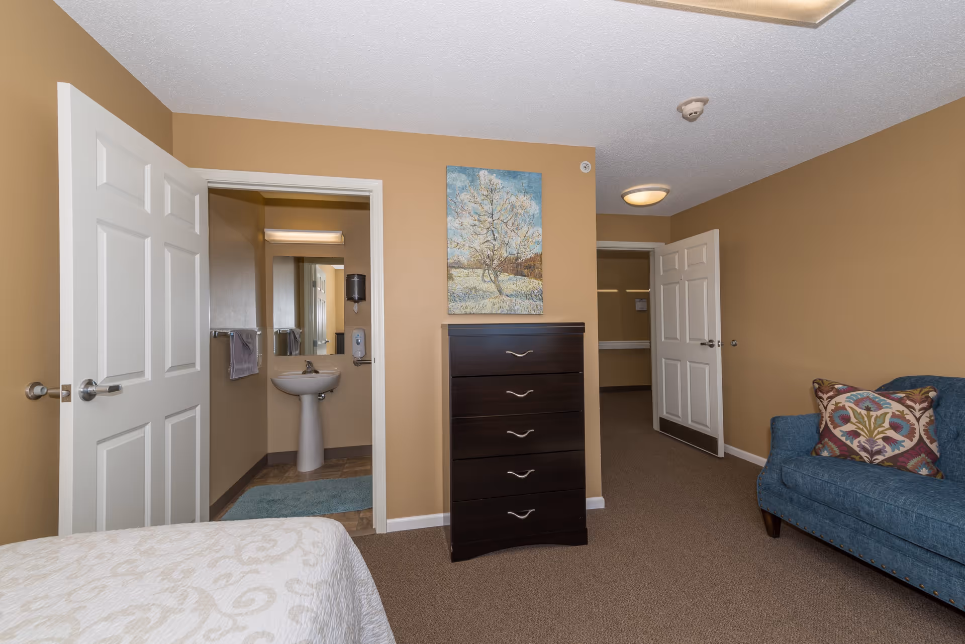 Interior view of a room in a senior living facility showing a bed with a white patterned bedspread, a dark wooden chest of drawers with a painting of a tree above it, an open door leading to a bathroom with a pedestal sink and mirror, and a blue upholstered couch with a decorative pillow.