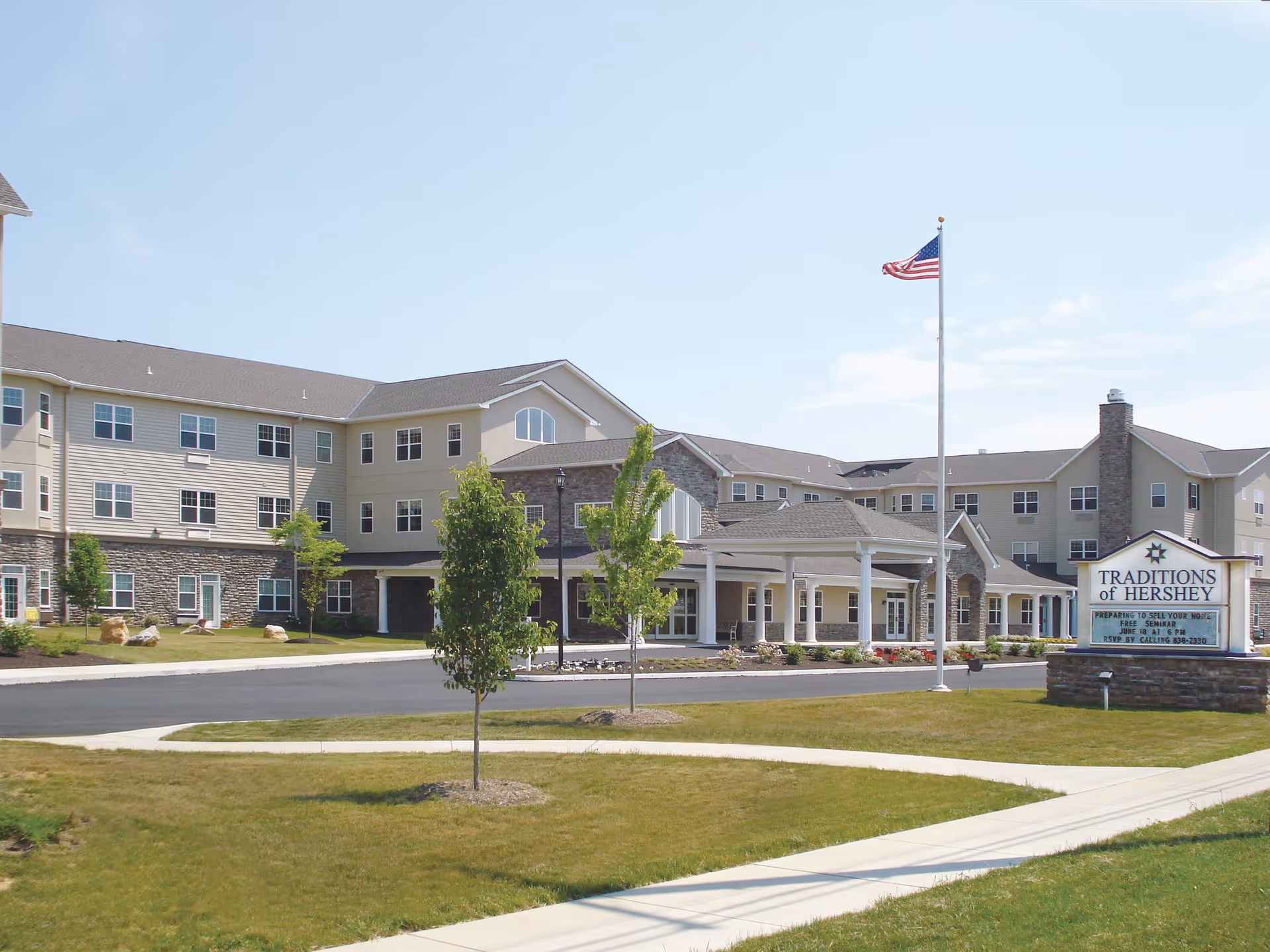 Exterior view of Traditions of Hershey, a large multi-story senior living facility with beige and stone facade, an American flag on a flagpole, a driveway, and a sign displaying the facility's name and event information.