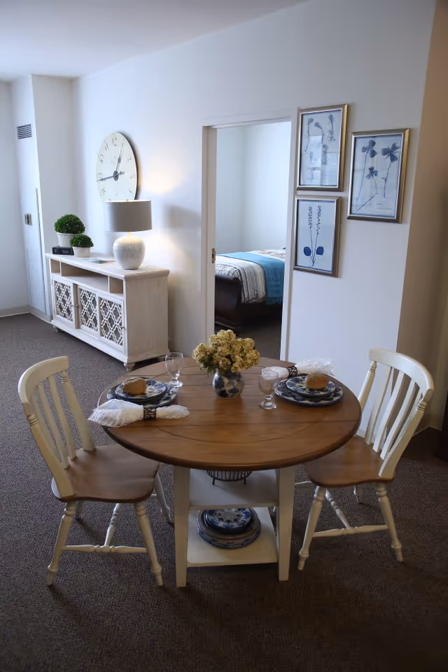 A small dining area with a round wooden table set for two, featuring plates, glasses, napkins, and a vase with yellow flowers. Two wooden chairs with white backs and legs are positioned at the table. In the background, there is a white sideboard with decorative plants and a large wall clock above it. A doorway leads to a bedroom with a bed covered in a striped blanket and a blue throw. Three framed botanical prints are hung on the wall next to the doorway.