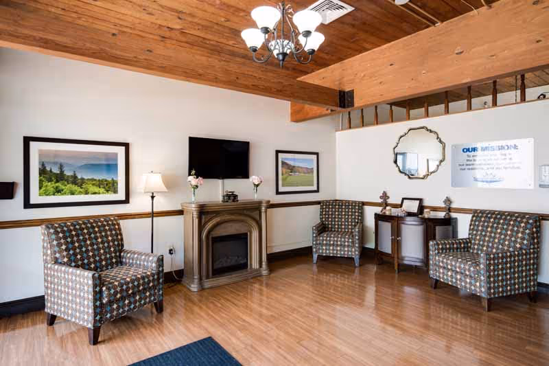 A cozy living room area with three patterned armchairs arranged around a small electric fireplace. Above the fireplace is a wall-mounted flat screen TV, and on either side of the fireplace are framed landscape photographs. The room has wooden flooring, a wooden ceiling with exposed beams, a chandelier, and a small side table with decorative items and a mirror on the wall.