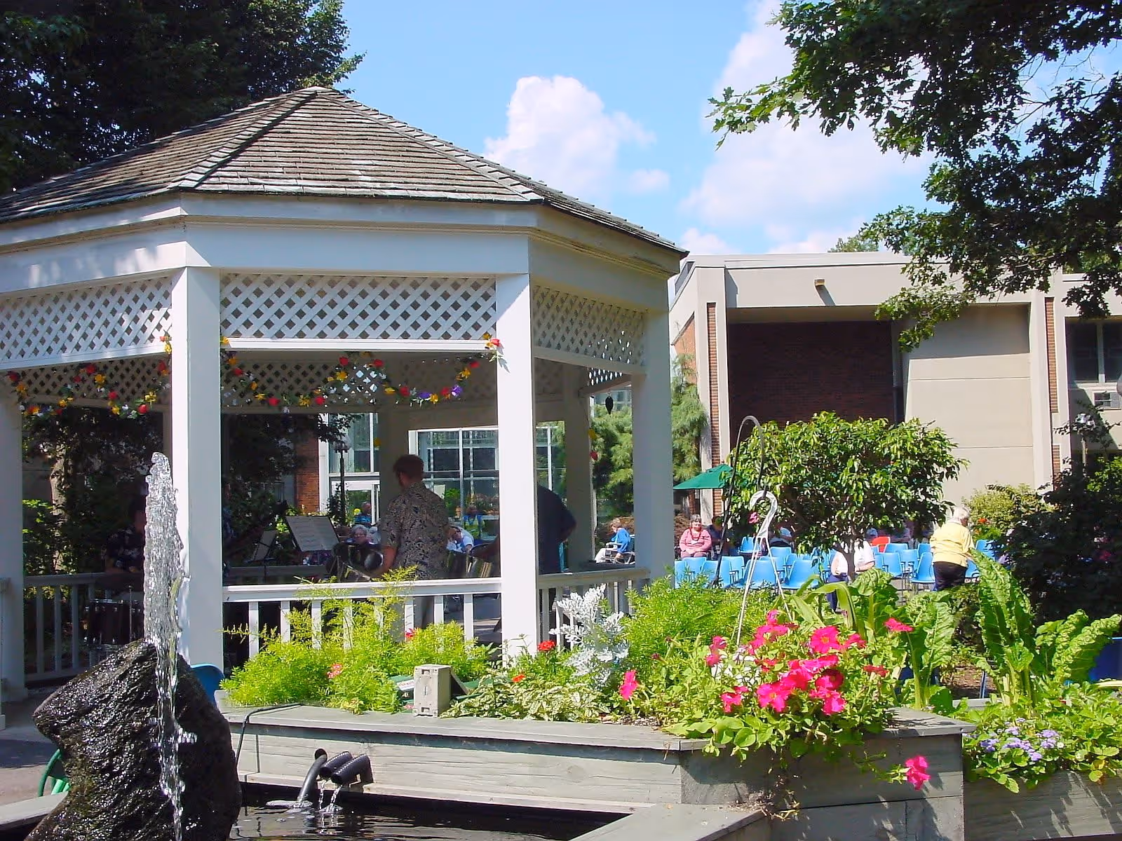Outdoor garden area at McLean a CCRC featuring a white gazebo decorated with flower garlands, a water fountain in the foreground, colorful flowers and greenery, and people seated on blue chairs in the background near a building under a sunny sky.