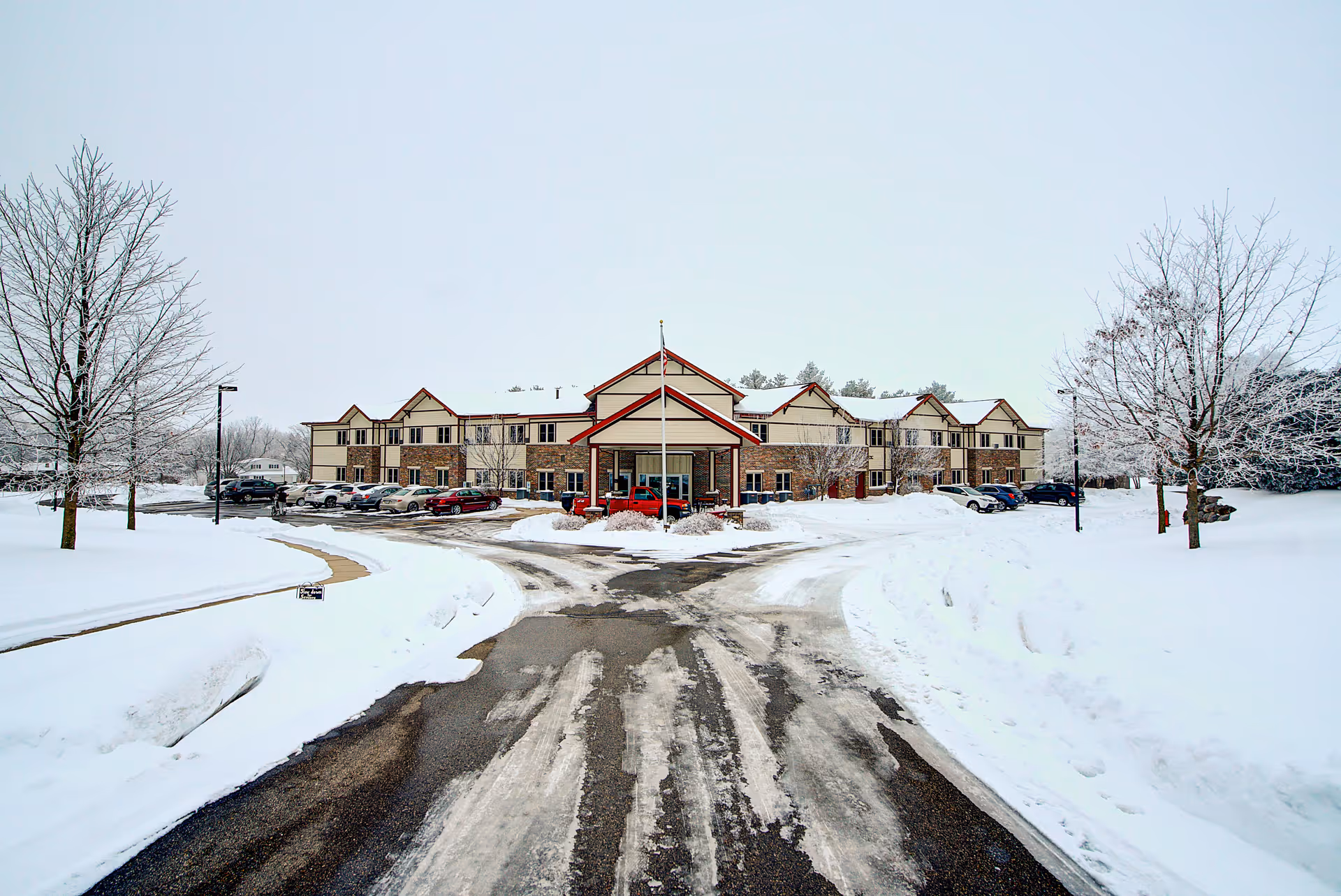 Front exterior view of The Heights at Evansville Manor building during winter with snow covering the ground, trees without leaves, a driveway leading to the entrance, and several parked cars.