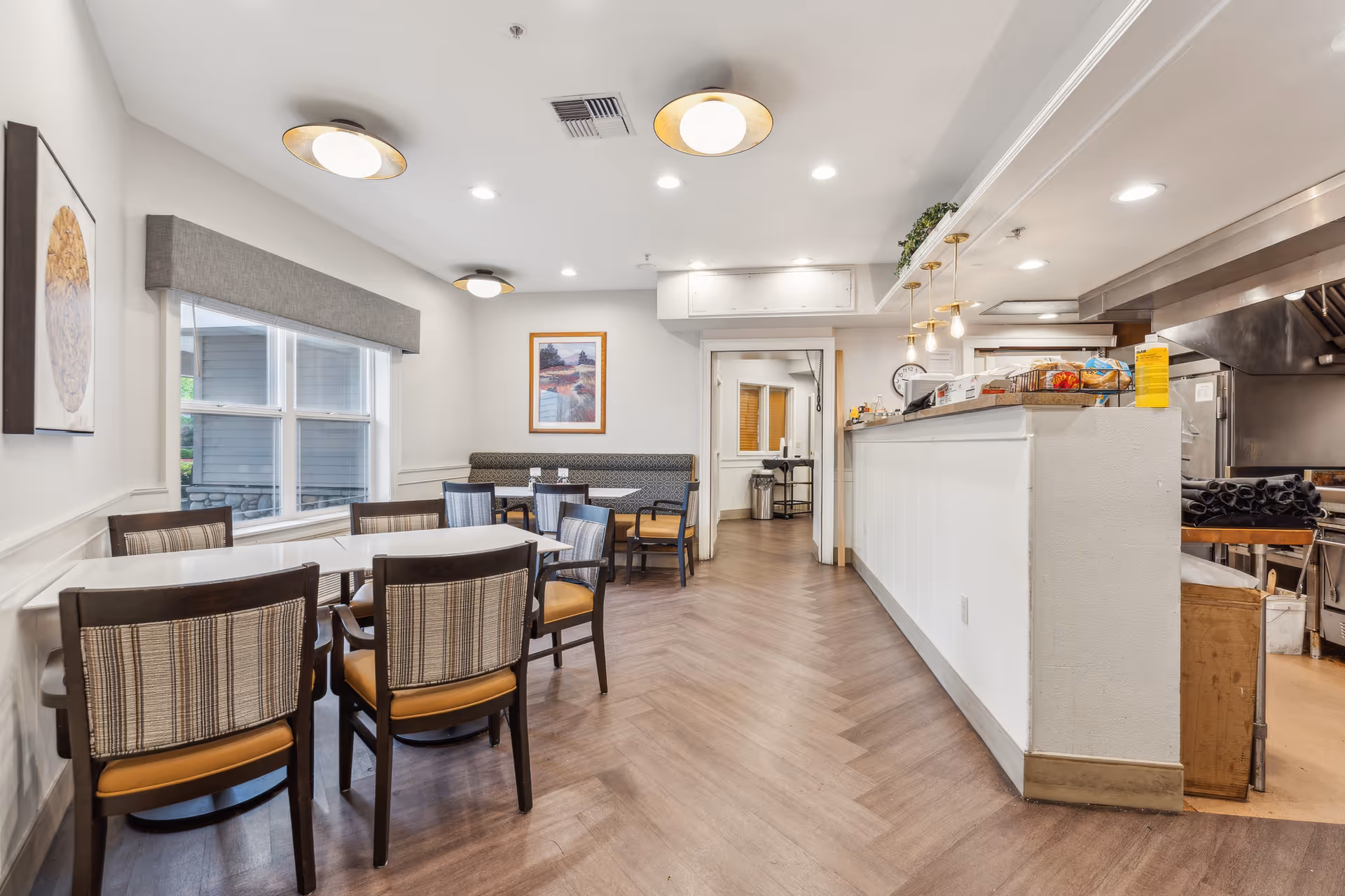 Interior view of a dining area in a senior living facility with several tables and chairs arranged neatly. The room features large windows with a gray valance, modern ceiling lights, framed artwork on the walls, and a visible kitchen area with a counter and hanging pendant lights.