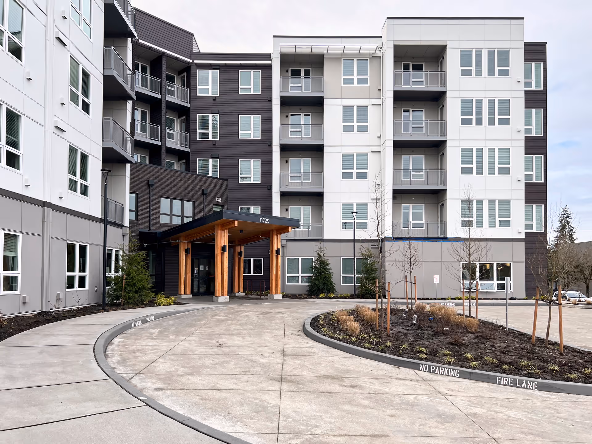 Exterior view of a modern multi-story senior living facility with a covered entrance supported by wooden pillars, surrounded by a curved driveway and landscaped area with young trees and plants.