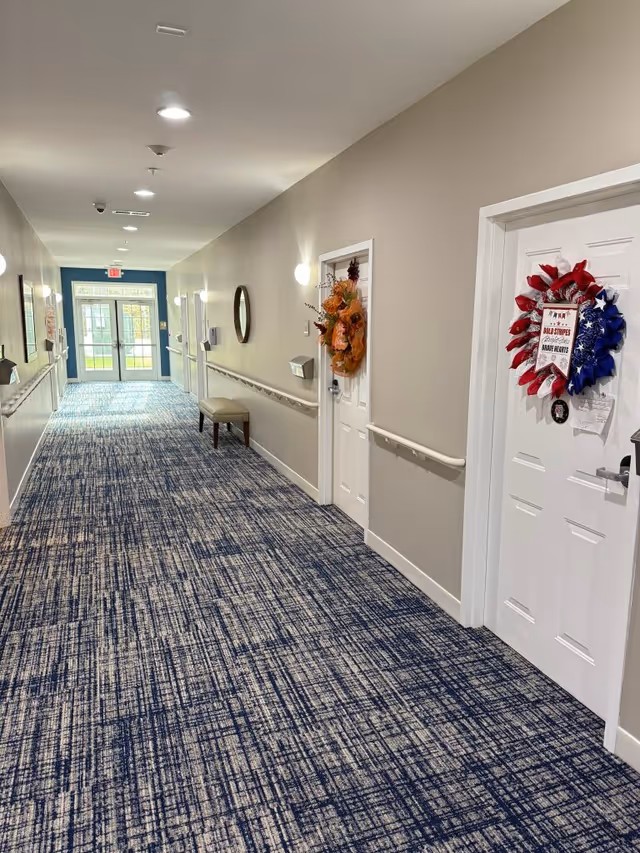 Well-lit interior hallway of an assisted living facility with patterned blue carpet, handrails, decorated resident doors, and exit doors at the far end.