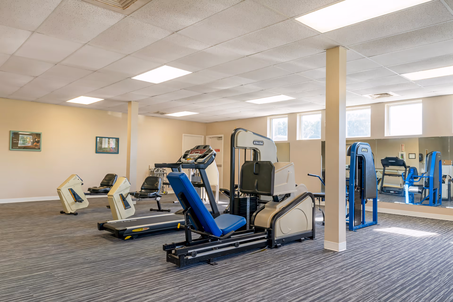 A rehabilitation gym room with various exercise equipment including a treadmill, leg press machine, and other fitness machines. The room has large windows letting in natural light, beige walls, and a carpeted floor. There are mirrors on one wall and two framed pictures on another wall.