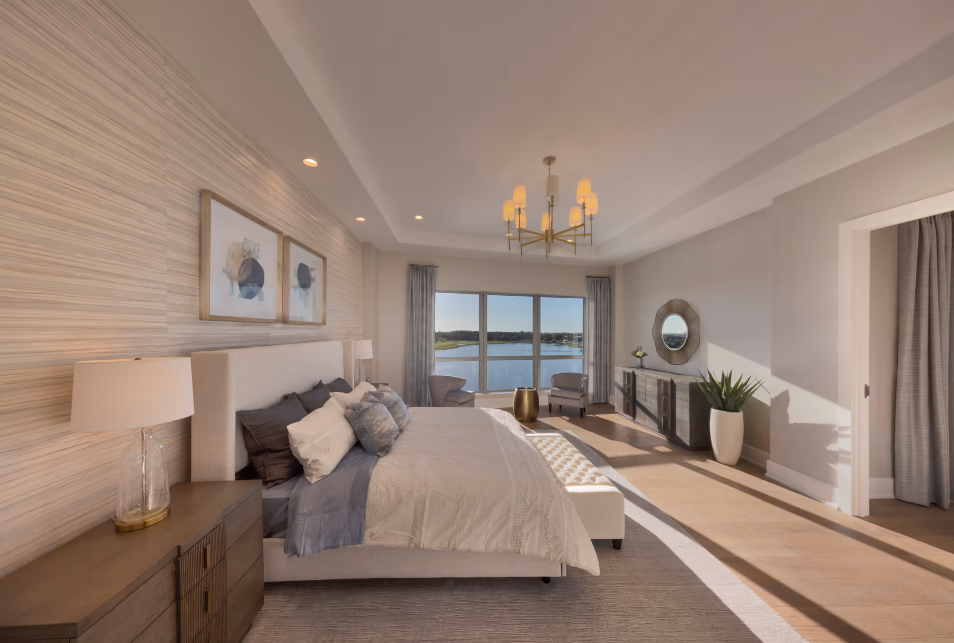 A spacious and bright bedroom with a large bed adorned with multiple pillows and a white comforter. The room features a wooden nightstand with a lamp, two framed abstract artworks above the bed, a chandelier hanging from the ceiling, and a large window offering a scenic view of a lake. There are two armchairs and a small round table near the window, a dresser with a round mirror, and a tall plant in a white pot. Sunlight streams into the room, creating a warm and inviting atmosphere.