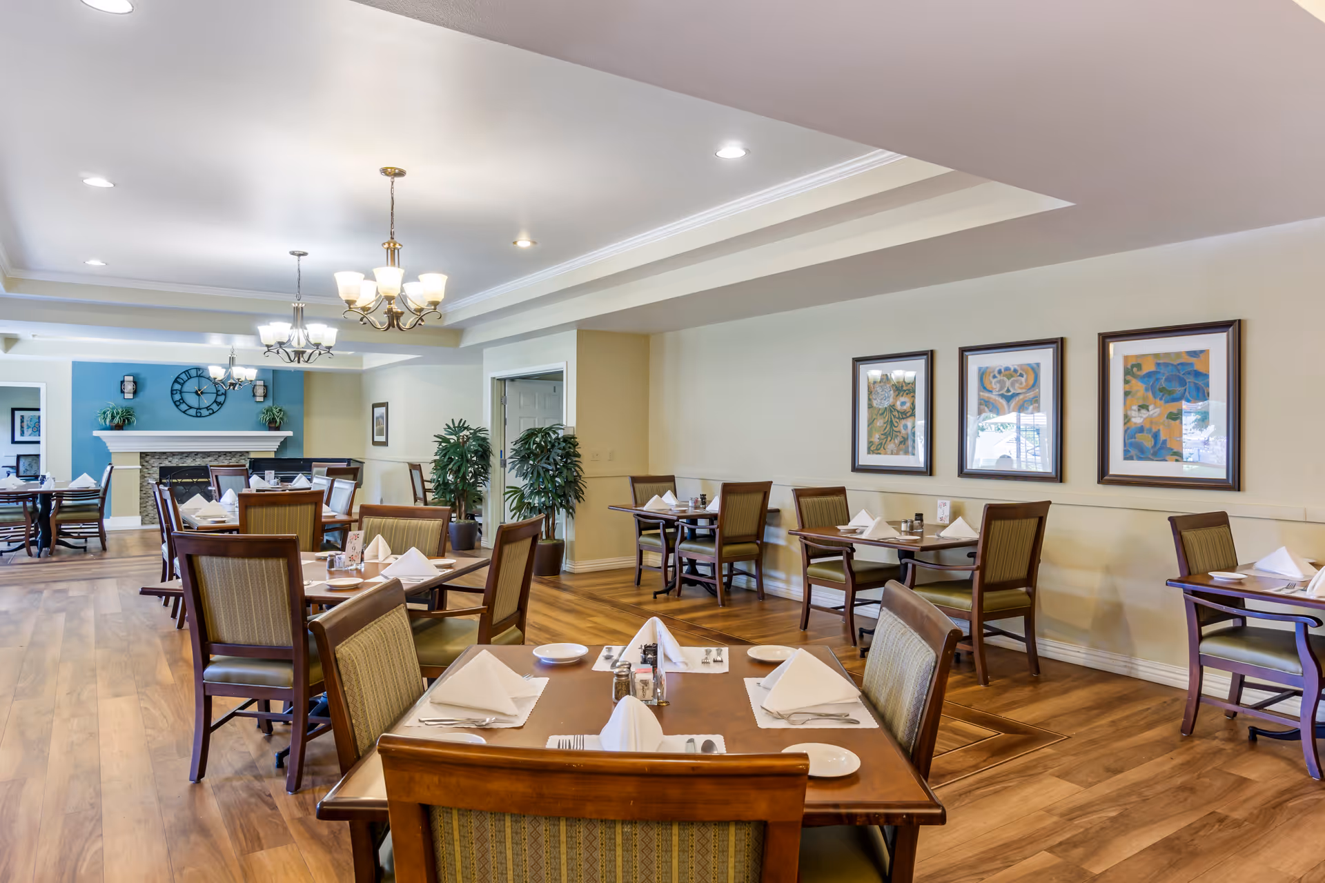 Spacious dining room with neatly set tables and chairs, chandeliers, framed artwork and hardwood floors.