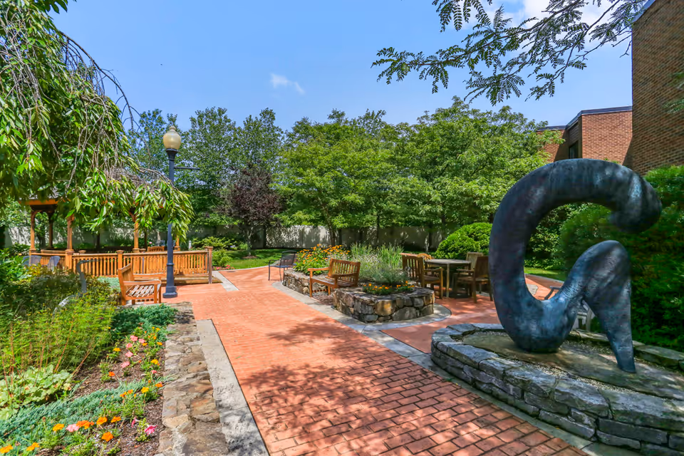 A sunny courtyard with brick walkways, wooden benches, a gazebo, landscaping, and a large abstract sculpture.
