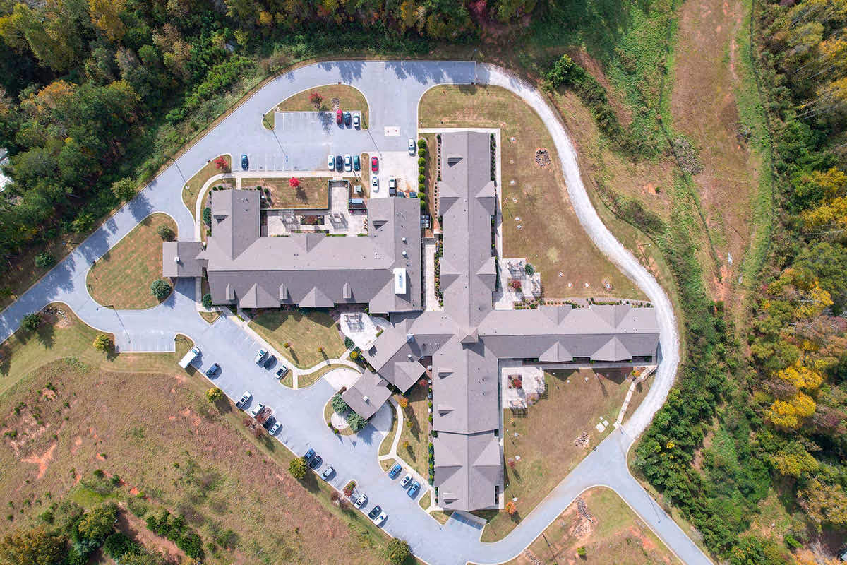 Aerial view of the Spring Park senior living facility showing a large building with multiple wings, surrounded by parking lots, driveways, and green landscaped areas with trees and grass.