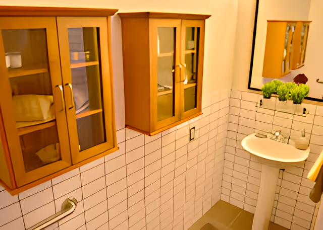 Small bathroom with a pedestal sink, wall-mounted glass-front cabinets, white tiled walls, a grab bar and a mirror with a potted plant.
