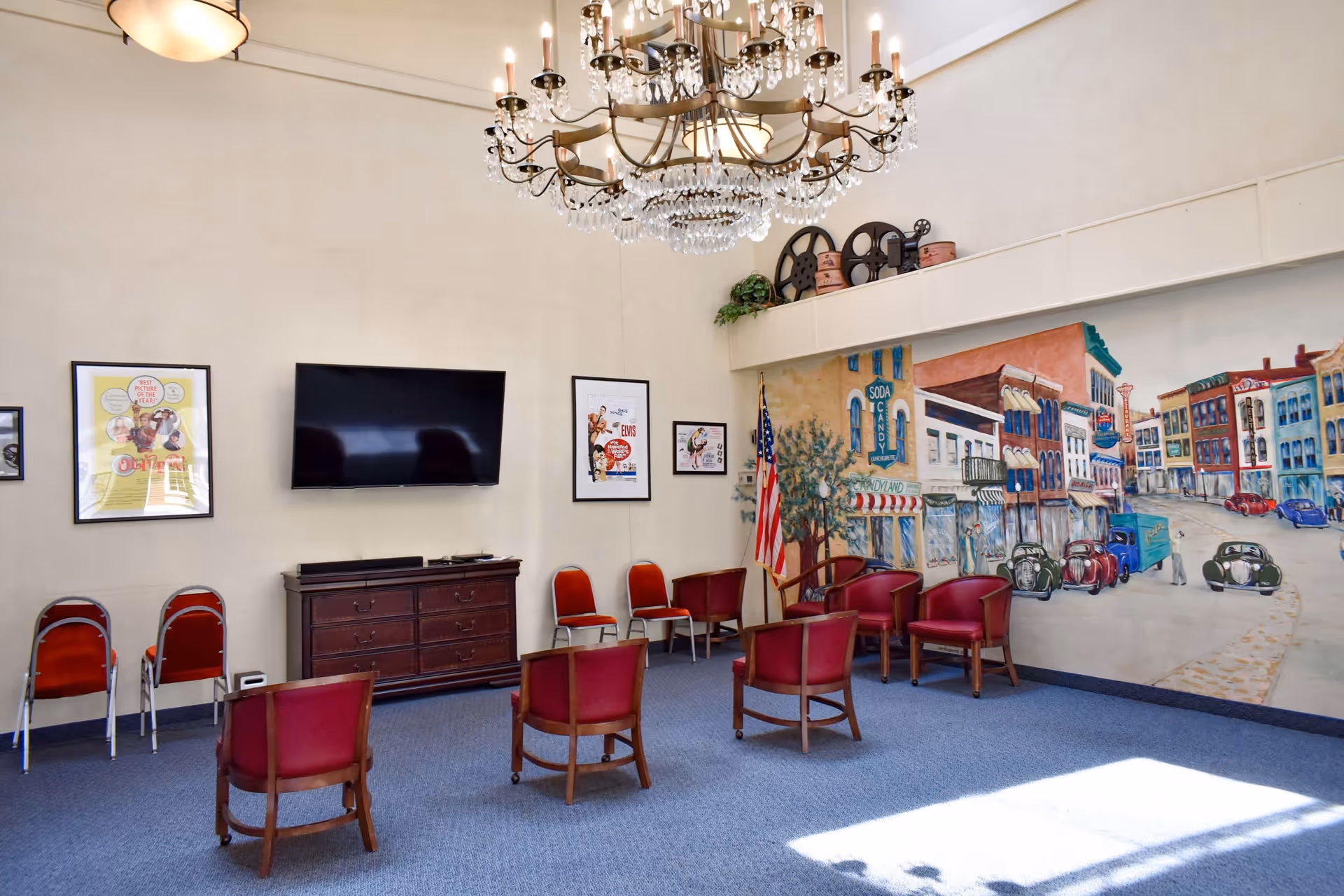 A spacious room with blue carpet and beige walls featuring a large mural of a vintage town street scene. The room has several red cushioned chairs arranged in a semi-circle, a dark wooden dresser with a flat-screen TV mounted above it, and framed posters on the walls. An American flag stands in the corner, and a large chandelier hangs from the ceiling.