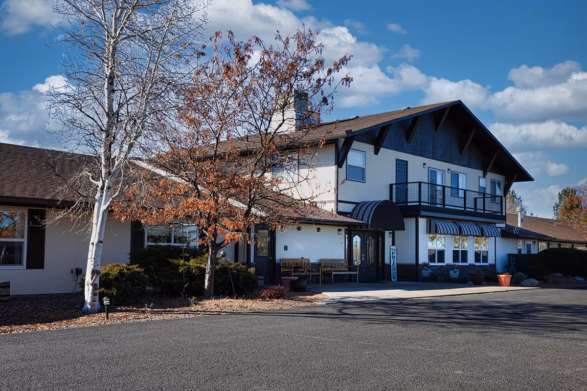 Front exterior of a two-story senior living building with a balcony, awnings, trees, and a paved driveway.