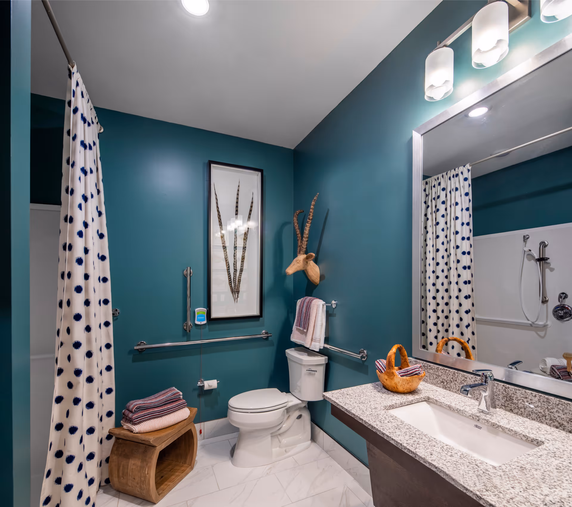 A modern bathroom with teal walls, a white toilet, a granite countertop with a sink, a large mirror, a shower with a white curtain featuring blue polka dots, and decorative elements including a framed picture and a wooden antelope head mounted on the wall.