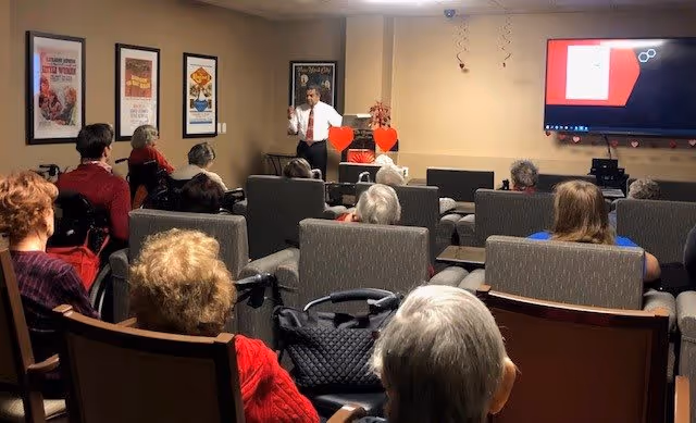 A group of elderly people seated in a room facing a man in a white shirt and red tie who is standing and speaking in front of them. The room has framed posters on the wall and a large screen displaying a presentation.