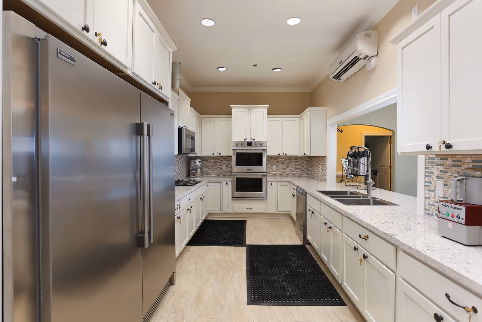 A modern kitchen with white cabinetry, stainless steel refrigerator, double wall ovens, microwave, dishwasher, and a sink with a high-arc faucet. The backsplash features a mosaic tile design, and there are two black floor mats on the light-colored tile floor. The kitchen is well-lit with recessed ceiling lights and has an air conditioning unit mounted on the wall.