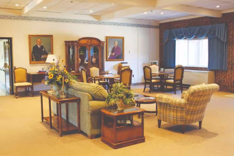 A spacious living room area in a senior living facility with beige carpet and cream-colored walls. The room features a green sofa and a plaid armchair arranged around wooden side tables with floral arrangements. In the background, there are several wooden dining tables with chairs, two framed portraits on the wall, a wooden cabinet, and a large window with blue drapes.