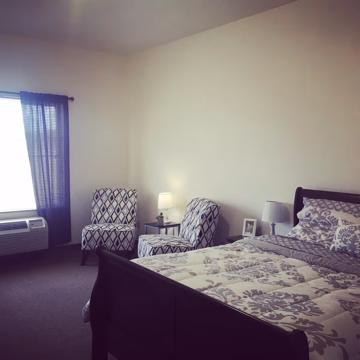 A bedroom with a bed featuring a patterned comforter and pillows, two patterned chairs, a small side table with a lamp, a window with dark curtains, and an air conditioning unit below the window.