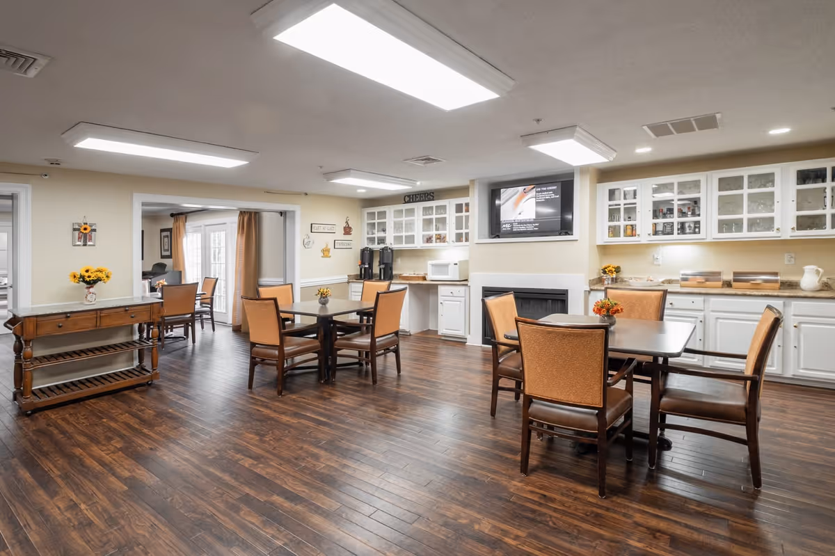 A spacious dining area with wooden floors, multiple tables and chairs, white cabinetry with glass doors, a microwave, coffee machines, and a wall-mounted TV above a fireplace. The room is well-lit with ceiling lights and decorated with small flower arrangements on the tables and a vase of sunflowers on a side table.