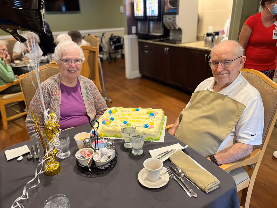 Two elderly residents sit at a dining table with a decorated sheet cake and place settings in a communal dining area.