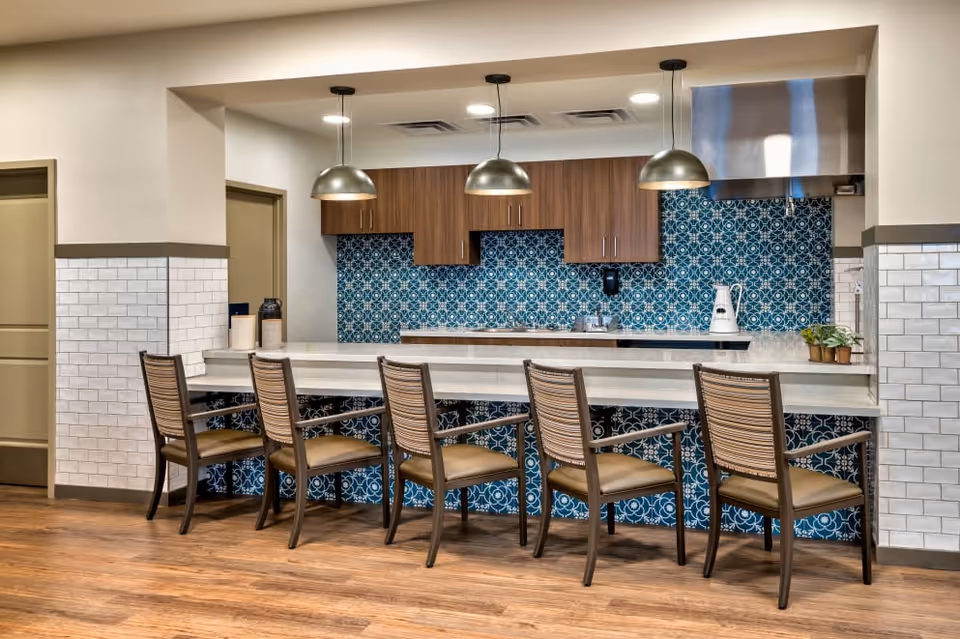 A modern kitchen area with a long white countertop and five wooden chairs with woven backs and tan cushions. The backsplash features a blue and white patterned tile design. Above the counter, there are three pendant lights with metallic shades. Wooden cabinets are mounted on the wall, and a stainless steel range hood is visible on the right side. The floor is wooden, and the walls are partially tiled with white subway tiles.