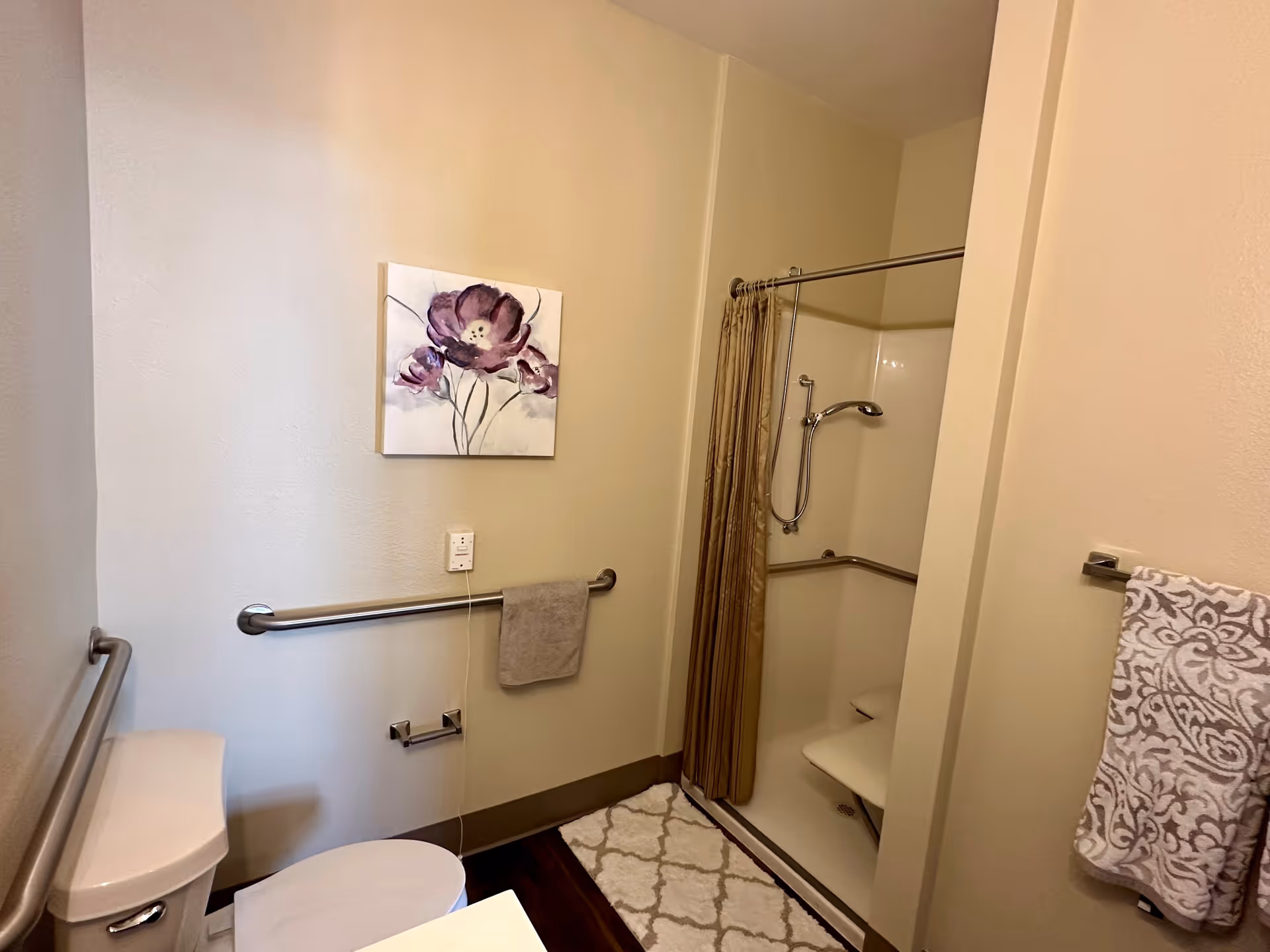 A bathroom with a toilet, a walk-in shower with a fold-down seat and grab bars, beige walls, a floral painting, and patterned towels hanging on towel racks.