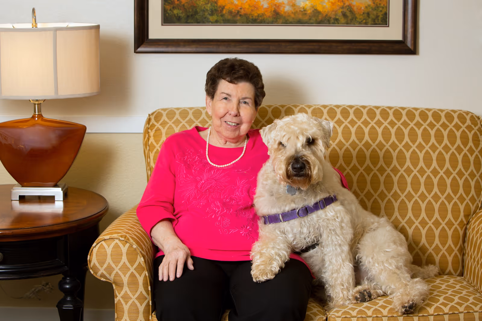 An elderly woman wearing a bright pink sweater and a pearl necklace sitting on a patterned yellow couch with a large light-colored dog wearing a purple harness sitting beside her. There is a wooden side table with a lamp on it to the left and a framed painting on the wall behind them.