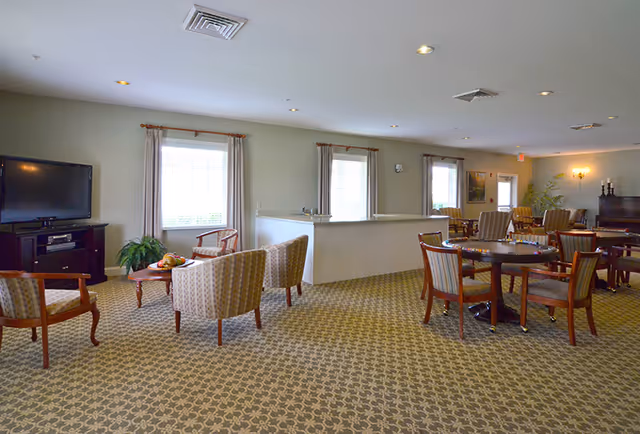 Spacious senior living common room with multiple seating areas, round tables and chairs, a TV on a stand and windows with curtains.